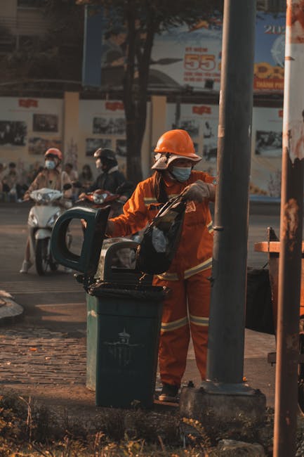 A worker dressed in an orange safety uniform and wearing a protective helmet and face mask is seen standing outdoors in an urban area near a grey metal pole. They are holding a black plastic bag and a