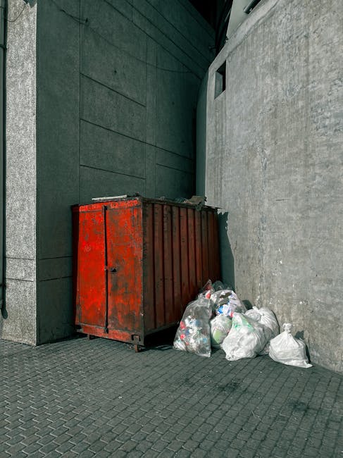 A worker dressed in an orange safety uniform and wearing a protective helmet and face mask is seen standing outdoors in an urban area near a grey metal pole. They are holding a black plastic bag and a