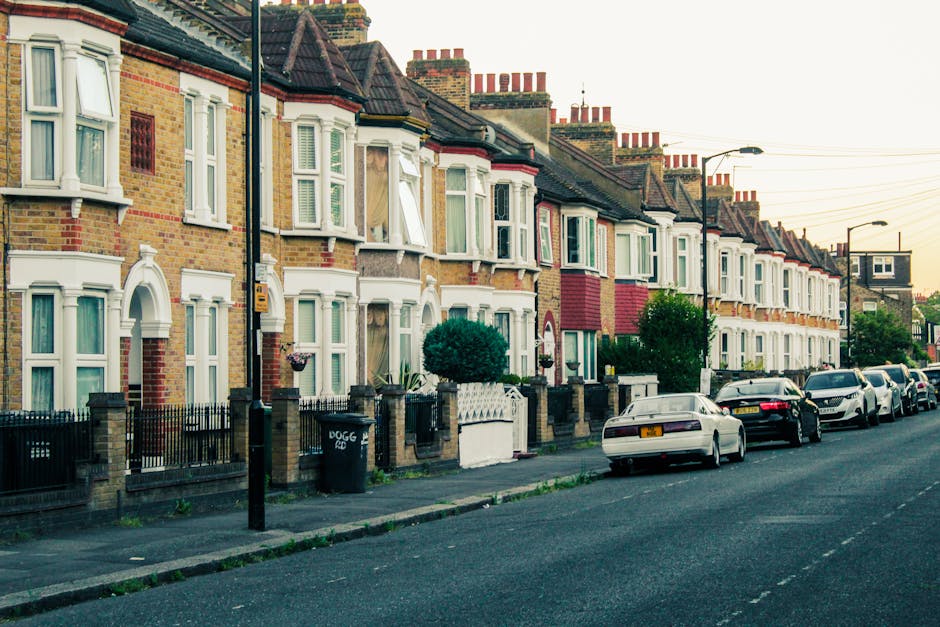 A row of traditional terraced houses with brick facades and bay windows, situated along a residential street under a clear sky during daytime. The houses feature various exterior finishes, including r